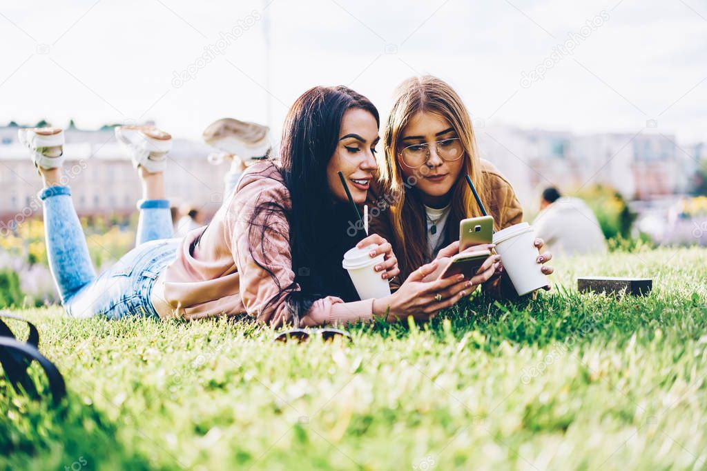 Two cheerful hipster girls lying on green grass while reading posts and watching videos in social networks on modern telephone devices spending leisure time together with tasty coffee outdoors in park