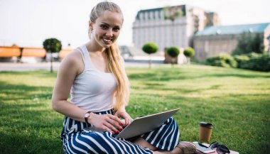 Sıradan giyinen neşeli kız öğrencinin portresi park çimlerinde öğreniyor bilgi ve eğitim için zaman ayırıyor, bilgisayarına gülümseyen bilgisayarlı mutlu kadın serbest çalışıyor.