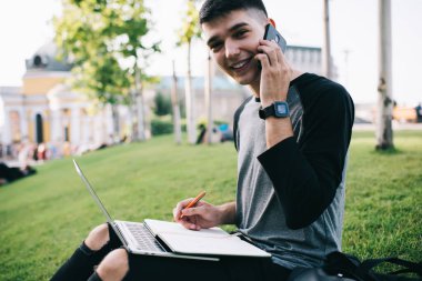 Bilgi kitapçığı ve modern dizüstü bilgisayarı olan neşeli erkek öğrencilerin portresi açık havada oturup danışmanlık için uluslararası sohbetler yapmak ve ders çalışma planı yaratmak için.