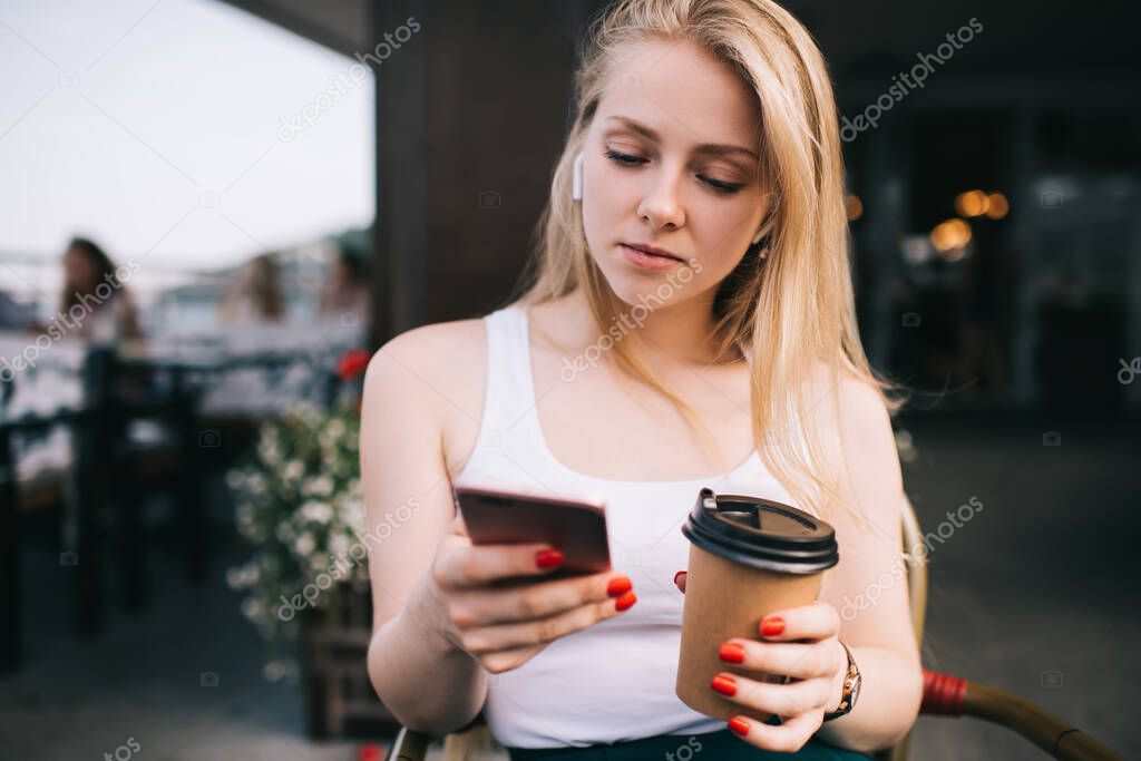 Millennial Caucasian woman in bluetooth headphones checking notification for stereo application sitting at street terrace with caffeine beverage in takeaway cup, hipster meloman opening sound playlist