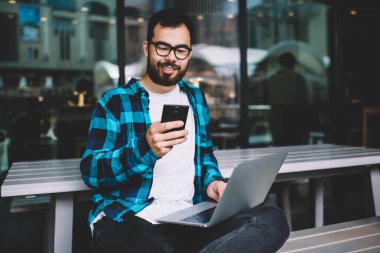 Genç serbest çalışan akıllı telefondaki uyarıyı uzaktan kontrol eden kablosuz ücretsiz internet kullanan hipster adam dizüstü bilgisayarda oturup sosyal ağlarda sohbet ediyor.