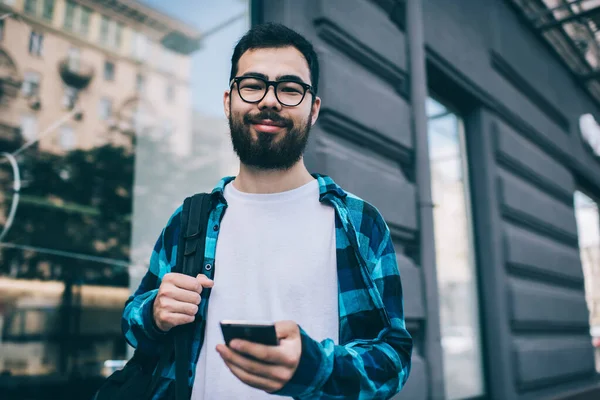 Half length portrait of positive millennial male travel blogger standing on urban setting and smiling at camera on leisure, happy hipster guy in spectacles for vision correction holding cellphone