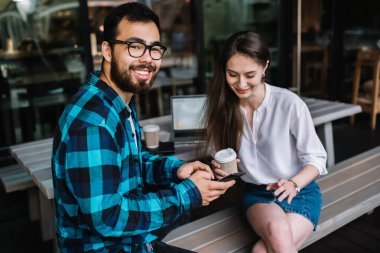 Birbirini seven pozitif çift internet sitesindeki yeni arkadaş paylaşımlarını okuyorlar. Modern akıllı telefon cihazında ücretsiz 4G internet bağlantısı kullanıyorlar. Açık havada kahve dükkanında oturuyorlar.