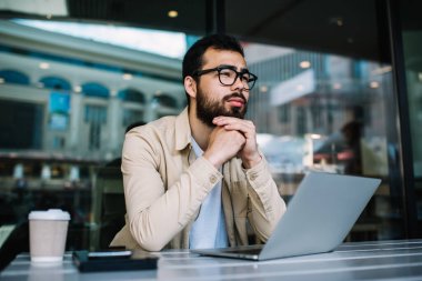 Gözlüklü düşünceli bir erkek programcı dizüstü bilgisayardaki veritabanının güncellenmesi üzerine kafa yoruyor kafeteryada elinde netbook ile oturmuş uygulamayla web sitesi oluşturma fikri üzerinde düşünüyor.
