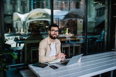 Görme düzeltmesi için optik gözlüklü mutlu Asyalı erkek öğrencinin portresi. Ders kitabında okul ödevi yaparken kameraya gülümsüyor ve bilgi araştırması yapmak için dizüstü bilgisayar kullanıyor.