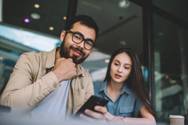 Gözlüklü neşeli sakallı hippi adamın portresi kameraya gülümsüyor ve güzel beyaz kız arkadaşıyla vakit geçirmekten zevk alıyor yanında oturuyor ve cep telefonunun ekranına bakıyor.