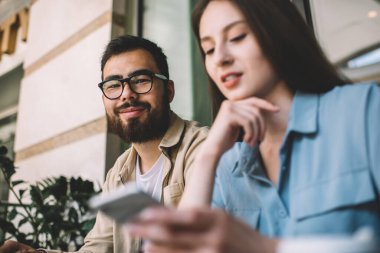 Optik gözlüklü gülümseyen hipster adamın portresi reklam alanında çekici kız arkadaşıyla birlikte oturuyor ve internette mesajlaşma yaptığı için canlı iletişimi görmezden geliyor.
