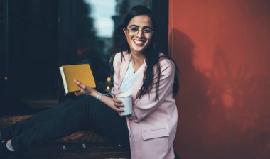 Kafenin yanında oturan, elinde kafein içeceği ve kameraya gülümseyen öğrenciler için web günlüğünün mutlu kadın yazarının portresi.