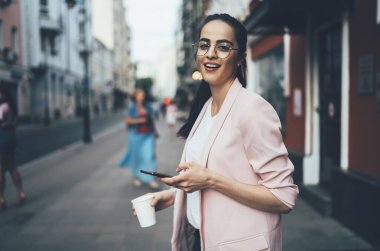 Gelişigüzel giyinen pozitif hippi kızın yarım boy portresi modern teknolojiyi telefon etmek ve kahve içmek için elinde tutarak şehir merkezini keşfederken kameraya gülümsüyor.