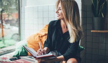 Neşeli beyaz kadın blogcu, sarı saçlı, pencereye bakıyor ve gülümsüyor. Heyecanlı bir fikirle kompozisyon hazırlıyor. Rahat bir kafede ders çalışmak için kendini iyi hissediyor.