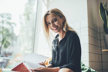Mükemmel beyaz dişleri olan neşeli bir kız öğrencinin portresi. Ders bölümlerini planlamak için kameraya gülümsüyor. Sıradan giyinen mutlu hippi kız evde eğitim için ders kitabı tutuyor.