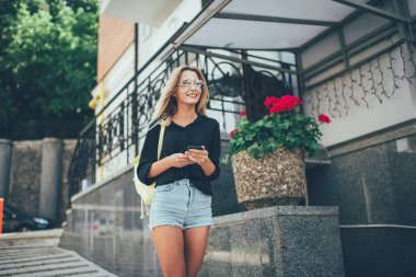 Gündelik giyinmiş neşeli kadın turist ellerinde modern akıllı telefon cihazları ve yaz tatillerinde şehir sokaklarında gezen şık güneş gözlüklü mutlu kadın dışarıda gülümsüyor.