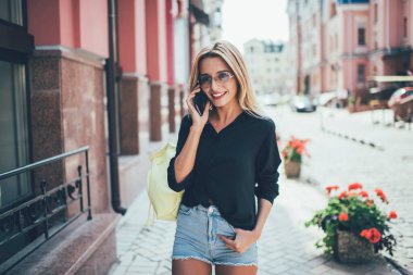 Modayı takip eden güneş gözlüklü kadın turistin yarım boy portresi. Akıllı telefon görüşmeleri sırasında gülümsüyor. Açık havada cep telefonuyla konuşan pozitif kadın.
