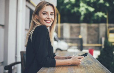 Teknoloji ve nesil kavramı, günlük tişört giymiş başarılı beyaz kadın blog yazarı portresi sokak kafeteryasında elinde dijital aletlerle kameraya gülümsüyor.