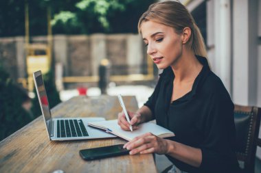 20 yaşındaki zeki beyaz kız öğrencinin öğrenim için dışarıda ders kitabı ve dizüstü bilgisayarla çalışmasının yan görüntüsü. Modern cep telefonu cihazında saati kontrol eden çekici bir kadın.