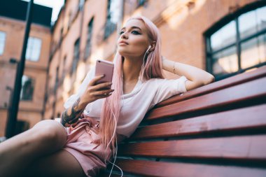 Kulaklık takan güzel genç bir kadın şehir bankında otururken podcast 'ten keyif alıyor. Pembe saçlı, cep telefonundan gelen ses kitabına odaklanmış beyaz bir meloman.