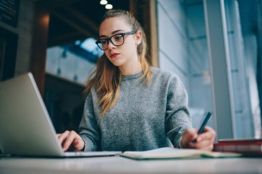 Kapalı alanlarda okuyan konsantre bir kız öğrenci netbook üzerinden okul ödevi için bilgi arıyor, gözlüklü hippi kız bilgisayarında ders kitabı okuyor.