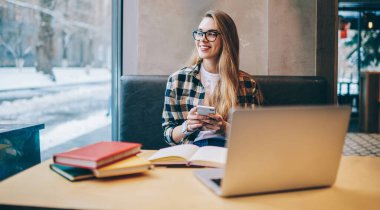 Gündelik giyinmiş, gülümseyen ve sınava hazırlanırken yüzünü çeviren modern teknoloji ve edebiyat kitaplarıyla dolu, elinde cep telefonu olan yeniyetme kız.