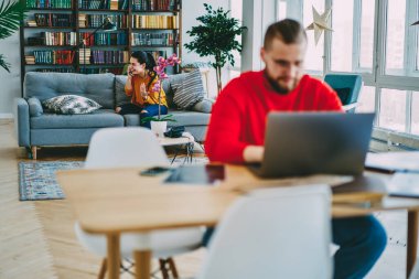 Gülümseyen beyaz bir kadın cep telefonuyla koltukta otururken kocası da şık bir şekilde tasarlanmış oturma odasının iç kısımlarında dizüstü bilgisayarda çalışıyor.