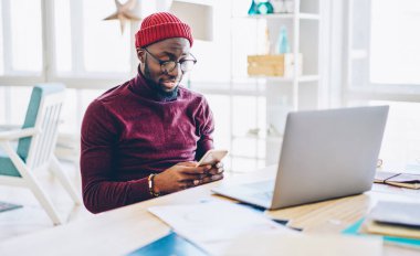 Gözlüklü Afro-Amerikan hipster adam cep telefonuyla mesaj gönderiyor evde internet üzerinden çalışırken, bin yıllık koyu tenli adam selülozdan yapılan işlemleri kontrol ediyor.