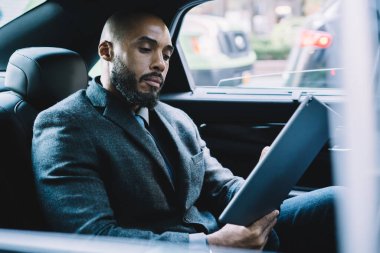 Koyu tenli erkek girişimci resmi giyinmiş. Borsacı web sitesinden finansal dünyadaki yenilikler hakkında e-posta almış. Havaalanına giden yolda bilet kesen adam.