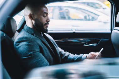 Resmi giyinmiş, koyu tenli, deneyimli bir patronun arka koltukta oturması ve akıllı telefon cihazında 4G kablosuz interneti kullanarak ortak e-postalarını kontrat şartlarıyla kontrol etmesi.