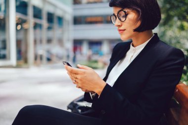 Görme düzeltmesi için optik gözlüklü konsantre bir kadın iş ortağına e-posta yazmak için mesaj yazıyor, genç kadın ekonomist dışarıda dinlenirken modern teknolojiyi sohbet için kullanıyor.