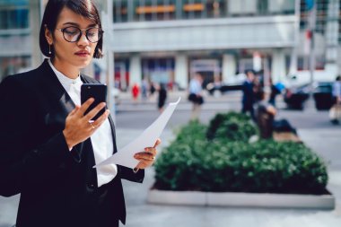 Resmi giysiler giymiş, gözlüklü bir iş kadını New York caddesinde elinde belgelerle ve modern akıllı telefon cihazıyla duruyor ve mali web sitesi ve evraklarda muhasebe kayıtlarını kontrol ediyor.