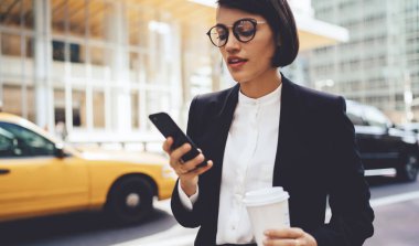 Şehir merkezinde kahve molası sırasında cep telefonuyla bankacılık için mobil uygulama kullanan kendine güvenen bir şirket sahibi gözlüklü kadın ekonomist SMS okurken paket servisi bardağını tutuyor.