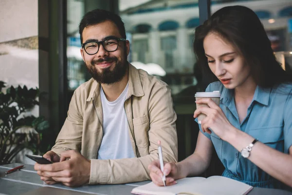 Görme düzeltmesi için optik gözlük takan neşeli erkek blog yazarı portresi kameraya gülümserken, yanında oturan ve okul sınavı için fikirler yazan bayan meslektaşıyla vakit geçiriyor.