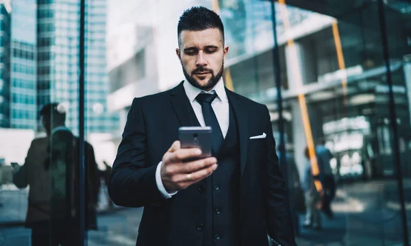 Serious executive manager 30 years old looking news on smartphone reading email about business using mobile phone with 4G internet near building in financial district, concept of online business