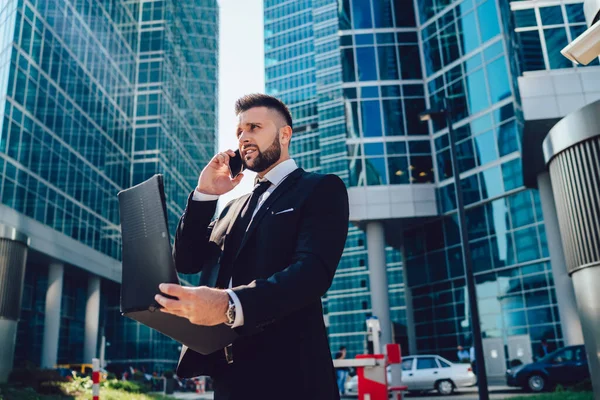 Puzzled male accounter with paper folder in hands checking information from report and calling to financial expert for discussing mistakes using roaming international connection in downtown city