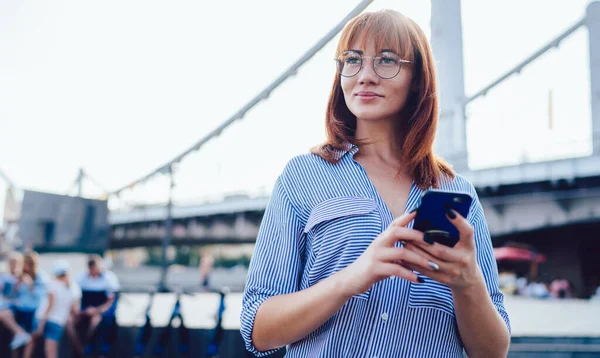 Beautiful millennial hipster girl holding modern mobile phone in hand and looking away while thinking about idea for new publication in social network, concept of technology and communication
