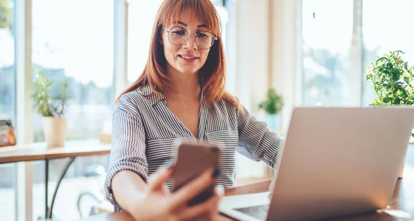 Mesafe mesafesinde cep telefonu cihazını kontrol eden olumlu kadın, akıllı telefon uygulamasını güncelleyen Kafkasyalı kadın bilgisayar başında oturuyor.
