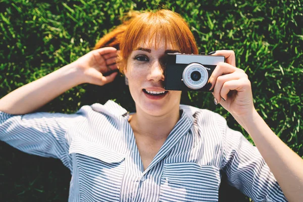 Top view of positive woman photographer in casual shirt lying on summer grass and making pictures via eyepiece on vintage camera, close up of happy female amateur enjoying time for favourite hobby