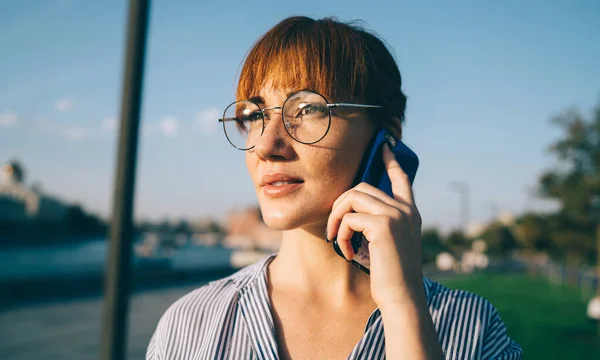 Millennial hipster girl in optical spectacles consultancy with operator about roaming connection listening useful information during smartphone conversation, concept of international communication