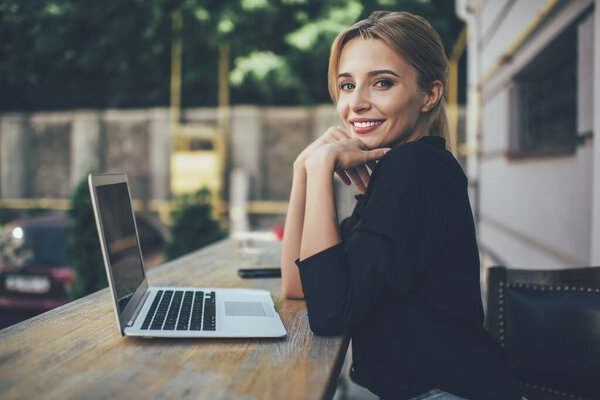 Portrait of sincere hipster girl skilled with laptop computer working remotely outdoors, happy Caucasian woman smiling at camera sitting at table with portable netbook, concept of millennial people