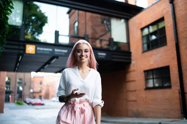 Half length portrait of cheerful millennial woman in spectacles for vision correction smiling at camera during free time in city, positive stylish hipster girl holding cellphone device and posing