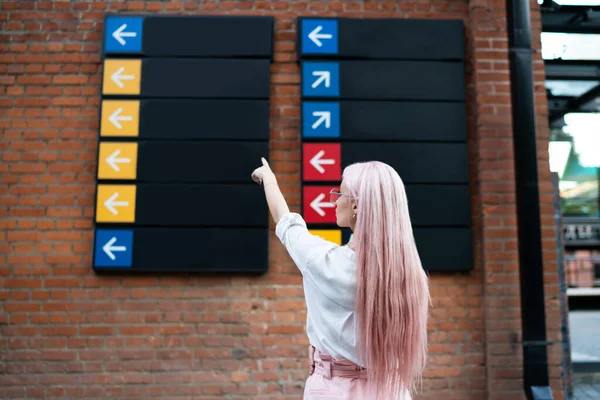 Uzun pembe saç stili, modaya uygun, modaya uygun, modaya uygun, bir sonraki durak olan turist bayanın şehir turizmini gösteren tabelayla duvara bakan şık hippi kızın arka planı.