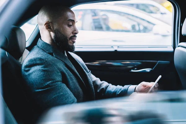 Resmi giyinmiş, koyu tenli, deneyimli bir patronun arka koltukta oturması ve akıllı telefon cihazında 4G kablosuz interneti kullanarak ortak e-postalarını kontrat şartlarıyla kontrol etmesi.