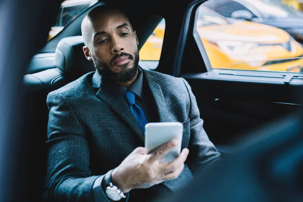 African American businessman using digital technology for development project during transportation in comfortable luxury car to office building, serious male expert sending sms with information