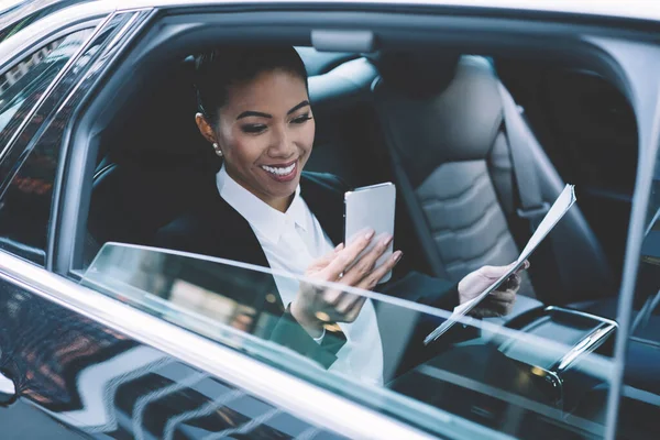 Cheerful woman in formal wear holding contract documents in hand and smiling from received email on modern cell smartphone device, happy female manager spending time for chatting on way to office