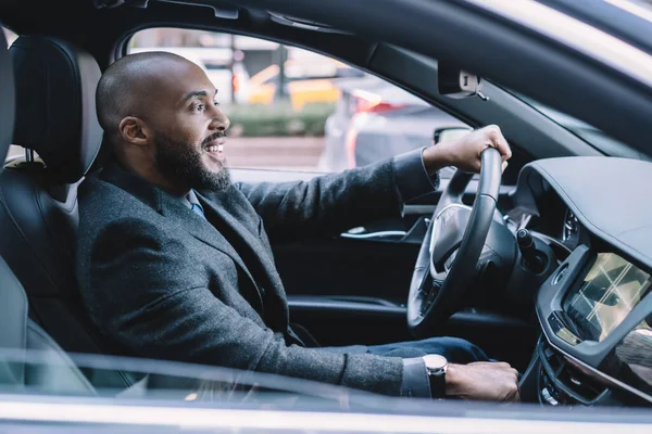 Side view of successful man in formal wear holding hand on steering wheel and enjoying driving getting to enterprise meeting in time, positive male entrepreneur smiling in new automobile car