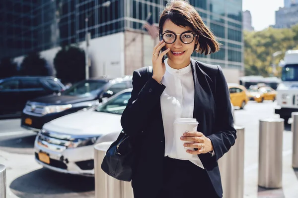 Half length portrait of positive female owner of company business enjoying coffee break in downtown and communication with partner via cellphone gadget, woman trader using smartphone device outdoors