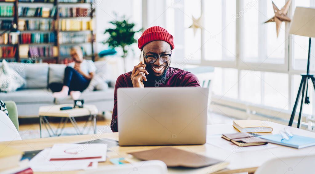 Young african american male happy about successful online job project talking with colleague on mobile phone and browse on laptop computer, hipster guy excited with winning online contest making cal