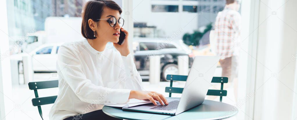 Young serious business woman in eyewear checking banking account on laptop while talking on cellular phone with service operator, female owner having mobile conversation while browsing information