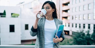 Eğitimle ilgili ders kitabı olan çekici bir kız öğrenci molada lezzetli kafein içeceği içiyor, resmi giyinmiş Türk kadın kahveyi seviyor ve düşünüyor.