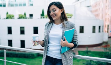 Eskiz defteriyle neşeli bir kız öğrenci ve elinde sıcak kafein bardağıyla geziyor. Teneffüste şehir ortamında gülüyor, dışarıda vakit geçirmek için kahveyle gülümsüyor.