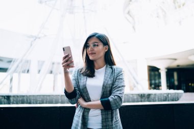 Gözlüklü olumlu kadın, modern cep telefonunda 4G kablosuz internete bağlı meslektaşlarıyla iletişim kurmak için yeni uygulama kuruyor, istatistikleri kontrol eden kadın blogcuları sorguluyor