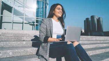 Modern bilgisayar cihazıyla şehir ortamında otururken mutlu bir metin yazarı gözlerini kaçırıyor ve gülümsüyor. Gözlüklü pozitif hippi kız dışarıda öğrenim görüp kendini iyi hissediyor.